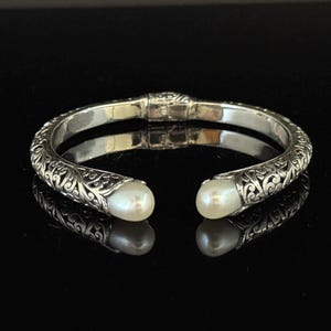 May include: A silver cuff bracelet with an open design, featuring intricate scrollwork and two white, pear-shaped pearls at the ends. The bracelet is displayed on a reflective black surface.