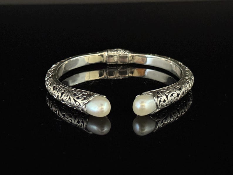 May include: A silver cuff bracelet with an open design, featuring intricate scrollwork and two white, pear-shaped pearls at the ends. The bracelet is displayed on a reflective black surface.