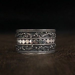 May include: A silver ring with a wide band featuring intricate floral designs and a row of small silver circles.