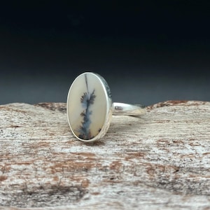 K&ouml;nnte beinhalten: Ovaler Ring mit Silberband. Der Ring hat einen cremefarbenen Stein mit einem schwarzen und grauen dendritischen Muster. Der Ring liegt auf einer Holzoberfl&auml;che.