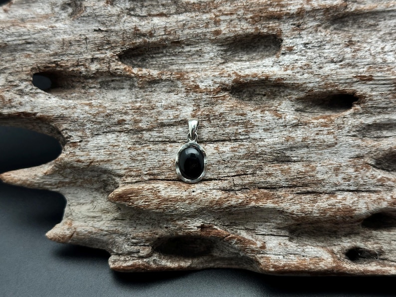 Puede incluir: Un colgante de plata con una piedra negra ovalada. El colgante se encuentra sobre un trozo de madera flotante desgastada. La madera tiene un aspecto natural y texturizado con varios agujeros e imperfecciones.