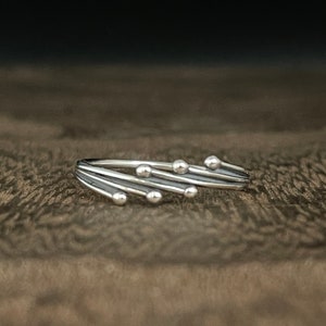 May include: A silver ring with a unique design featuring multiple thin bands with small round beads. The ring is on a brown textured surface.