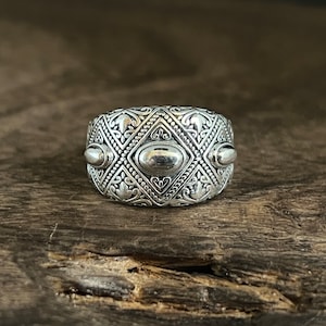 Pode incluir: Um anel de prata detalhado com um design ornamentado. O anel apresenta um padrão intrincado de diamantes, motivos florais e detalhes ovais. O anel é exibido em uma superfície de madeira rústica, destacando seu artesanato e estilo.
