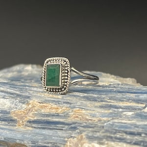 Puede incluir: Un anillo de plata con una piedra preciosa verde engastada en un bisel cuadrado. El anillo tiene una banda delicada y retorcida.
