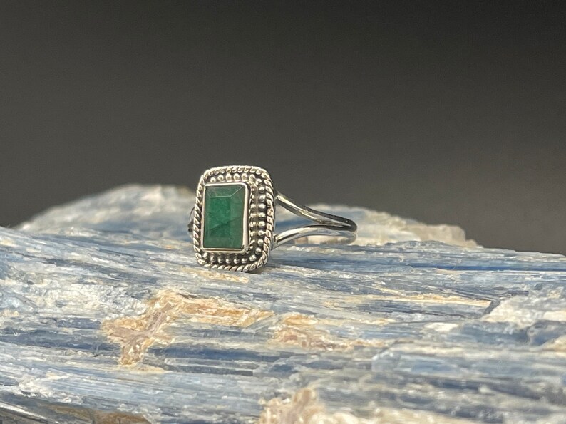Puede incluir: Un anillo de plata con una piedra preciosa verde engastada en un bisel cuadrado. El anillo tiene una banda delicada y retorcida.