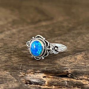 Könnte beinhalten: Ein Silberring mit einem ovalen, blauen Opal-Cabochon in einer dekorativen Fassung. Der Metallring ist mit einem gedrehten Seilmuster um den Stein herum sowie kleinen blattartigen Gravuren an den Seiten der Ringschiene verziert.