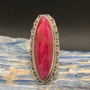 May include: A large, oval-shaped ring featuring a vibrant, faceted, ruby-red gemstone. The gemstone is set in a silver-toned metal with an ornate, filigree-style border. The ring's design is symmetrical and eye-catching, suitable for a statement piece.