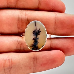 K&ouml;nnte beinhalten: Silberring mit einem ovalen Dendritachat-Stein. Der Stein hat einen cremefarbenen Hintergrund mit schwarzen und dunkelblauen, baumartigen Mustern. Der Ring ist in einem Silberband gefasst.