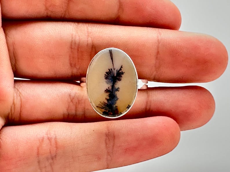 K&ouml;nnte beinhalten: Silberring mit einem ovalen Dendritachat-Stein. Der Stein hat einen cremefarbenen Hintergrund mit schwarzen und dunkelblauen, baumartigen Mustern. Der Ring ist in einem Silberband gefasst.