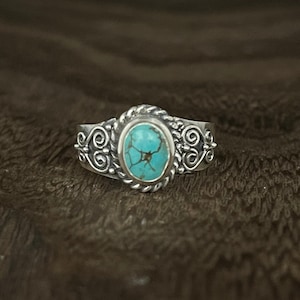 Op de afbeelding: Een zilveren ring met een turquoise steen in een decoratieve zetting. De band is versierd met ingewikkelde krulwerk.