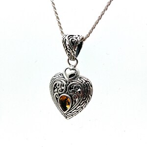 Puede incluir: Un collar con un colgante en forma de coraz&oacute;n de plata, adornado con un peque&ntilde;o dije en forma de coraz&oacute;n y una piedra preciosa amarilla en el centro. El colgante presenta intrincados detalles de filigrana.