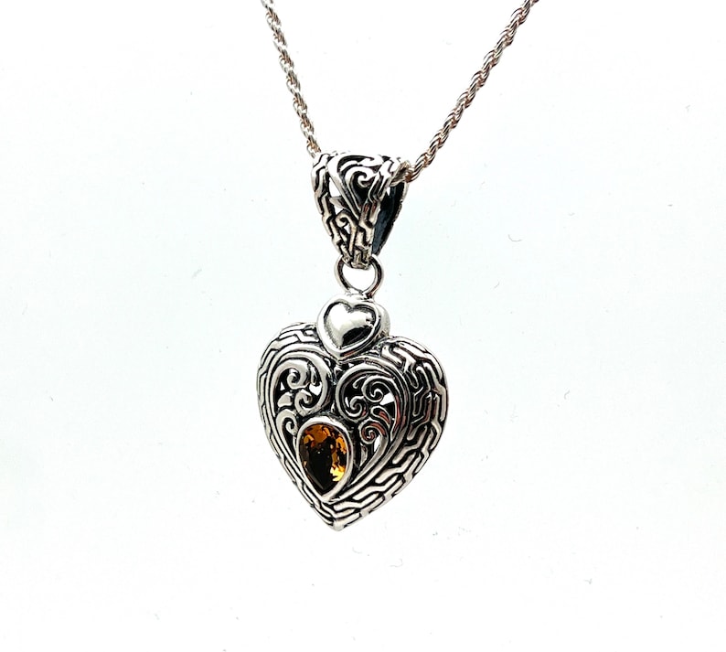 Puede incluir: Un collar con un colgante en forma de coraz&oacute;n de plata, adornado con un peque&ntilde;o dije en forma de coraz&oacute;n y una piedra preciosa amarilla en el centro. El colgante presenta intrincados detalles de filigrana.