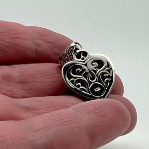 May include: A silver heart-shaped pendant with an intricate filigree design. The pendant has a small round opening in the center.