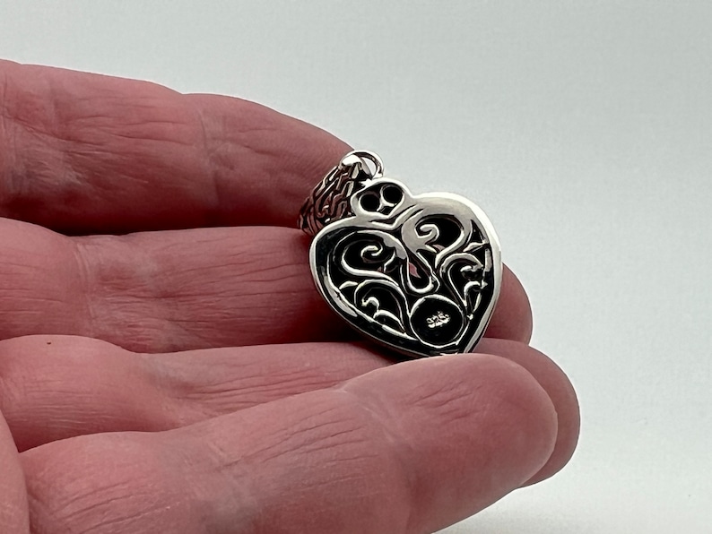 May include: A silver heart-shaped pendant with an intricate filigree design. The pendant has a small round opening in the center.