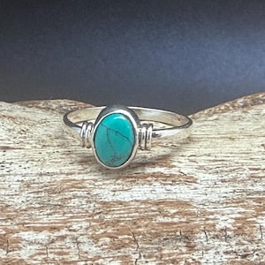 Op de afbeelding: Een zilveren ring met een ovale turkoois steen. De ring heeft een eenvoudige band en decoratieve accenten aan weerszijden van de steen. De turkoois steen heeft een levendige blauwgroene kleur.