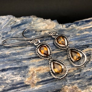 May include: A pair of silver drop earrings featuring two faceted, pear-shaped amber-colored gemstones set within twisted metal frames. Each earring is suspended from a simple silver hook, showcasing a vertical design with two stones per earring.