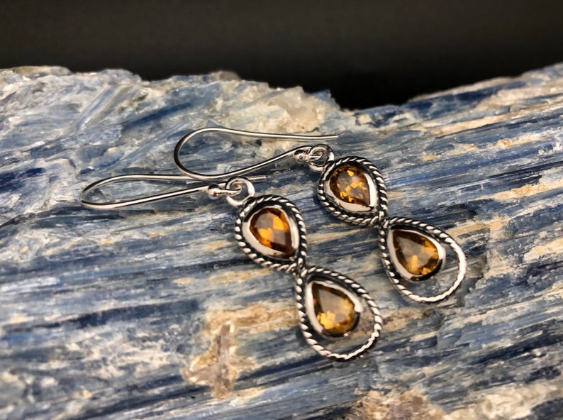 May include: A pair of silver drop earrings featuring two faceted, pear-shaped amber-colored gemstones set within twisted metal frames. Each earring is suspended from a simple silver hook, showcasing a vertical design with two stones per earring.