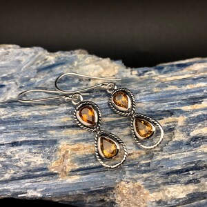 May include: A pair of drop earrings featuring two teardrop-shaped amber-colored stones set in twisted silver metal frames. Each earring has a hook-style closure and a vertical design with one stone positioned above the other in a linked arrangement.