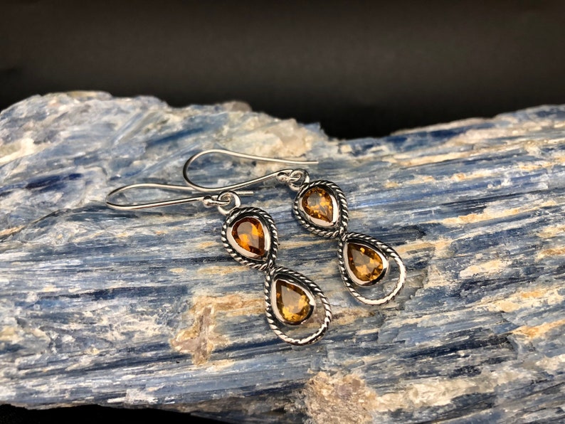 May include: A pair of drop earrings featuring two teardrop-shaped amber-colored stones set in twisted silver metal frames. Each earring has a hook-style closure and a vertical design with one stone positioned above the other in a linked arrangement.