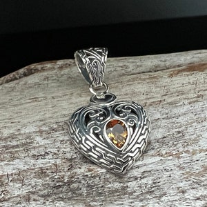 Puede incluir: Un colgante de plata en forma de coraz&oacute;n con un dise&ntilde;o intrincado y una piedra preciosa marr&oacute;n en el centro.