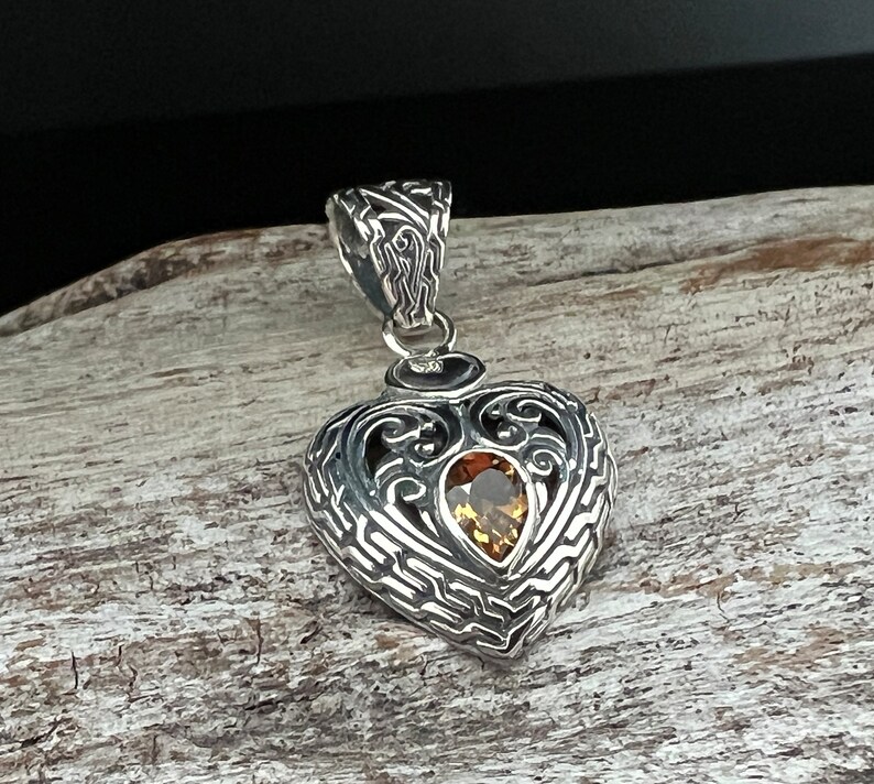 Puede incluir: Un colgante de plata en forma de coraz&oacute;n con un dise&ntilde;o intrincado y una piedra preciosa marr&oacute;n en el centro.
