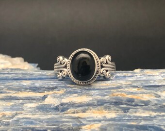 May include: A silver ring featuring an oval black gemstone set in an ornate bezel. The ring has a detailed design with scrolls and beading.