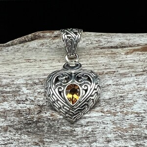 Puede incluir: Un colgante de plata en forma de coraz&oacute;n con un intrincado dise&ntilde;o de filigrana y una piedra preciosa amarilla en el centro.