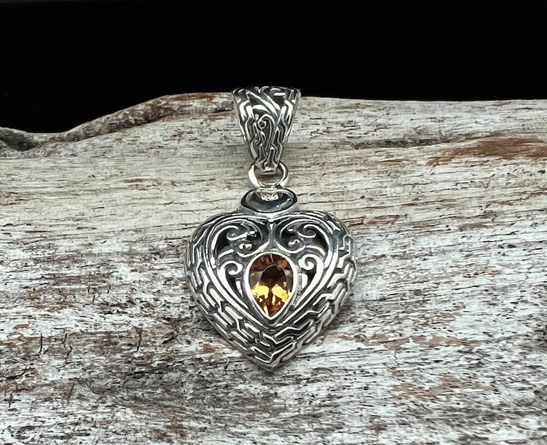 Puede incluir: Un colgante de plata en forma de coraz&oacute;n con un intrincado dise&ntilde;o de filigrana y una piedra preciosa amarilla en el centro.