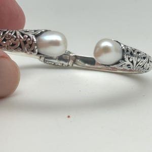 May include: A silver-colored open cuff bracelet with an ornate, filigree design. The bracelet features two large, round, white pearls. The bracelet is held by a hand against a white background.