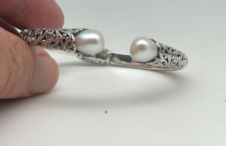 May include: A silver-colored open cuff bracelet with an ornate, filigree design. The bracelet features two large, round, white pearls. The bracelet is held by a hand against a white background.