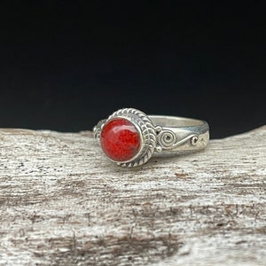 May include: A silver ring featuring a round, vibrant red stone. The stone is set in a decorative bezel with a rope-like border. The band has a swirling design on the sides, creating an elegant and eye-catching piece of jewelry.