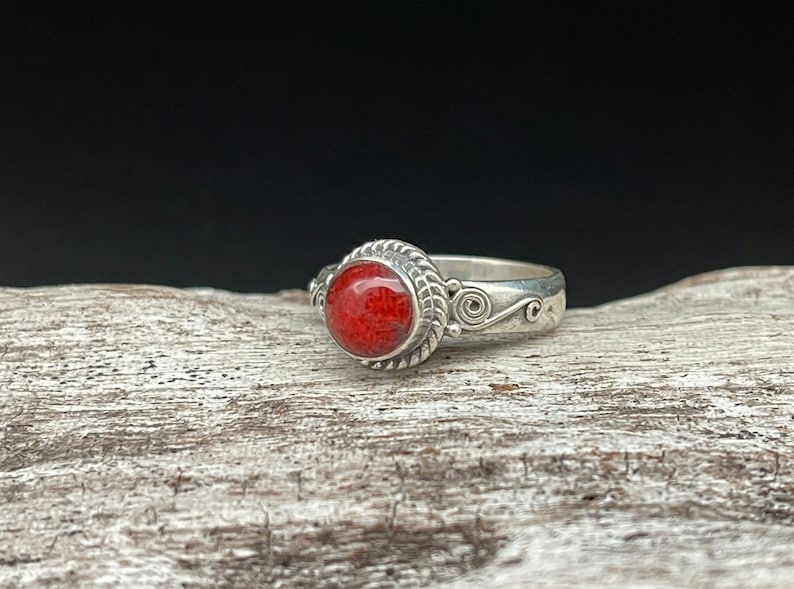 May include: A silver ring featuring a round, vibrant red stone. The stone is set in a decorative bezel with a rope-like border. The band has a swirling design on the sides, creating an elegant and eye-catching piece of jewelry.