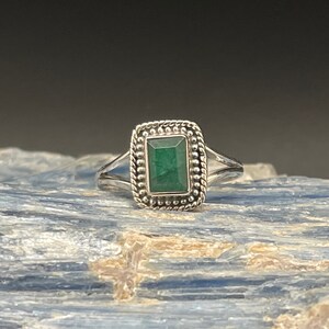 Puede incluir: Un anillo de plata con una esmeralda verde engastada en un bisel cuadrado. El anillo tiene un borde con cuentas y una banda retorcida.