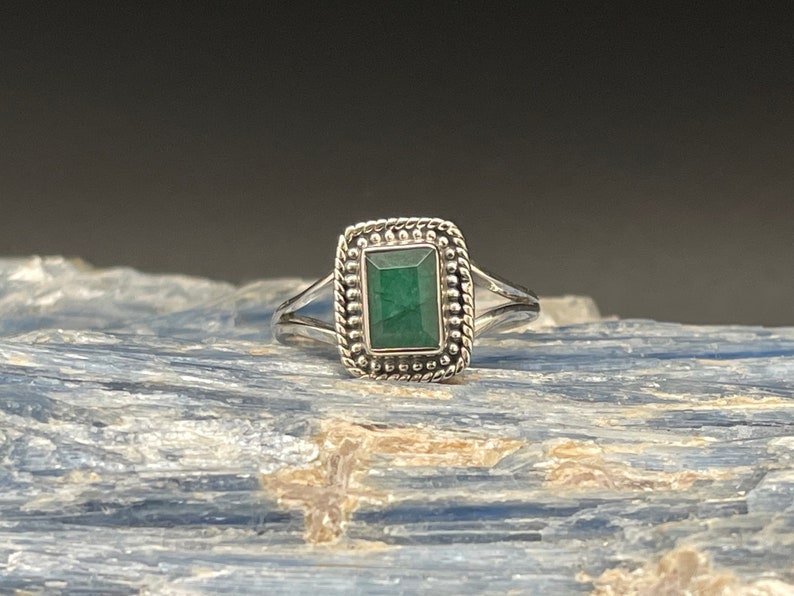 Puede incluir: Un anillo de plata con una esmeralda verde engastada en un bisel cuadrado. El anillo tiene un borde con cuentas y una banda retorcida.