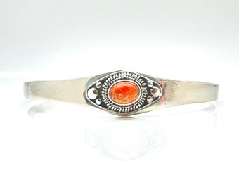 May include: A silver bracelet with a decorative oval-shaped design featuring a bright orange stone.