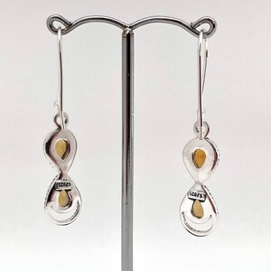 May include: A pair of silver drop earrings featuring two stacked, teardrop-shaped settings with light yellow stones. Each earring has a hook wire and a small rectangular plaque stamped with the text 925, indicating sterling silver composition.