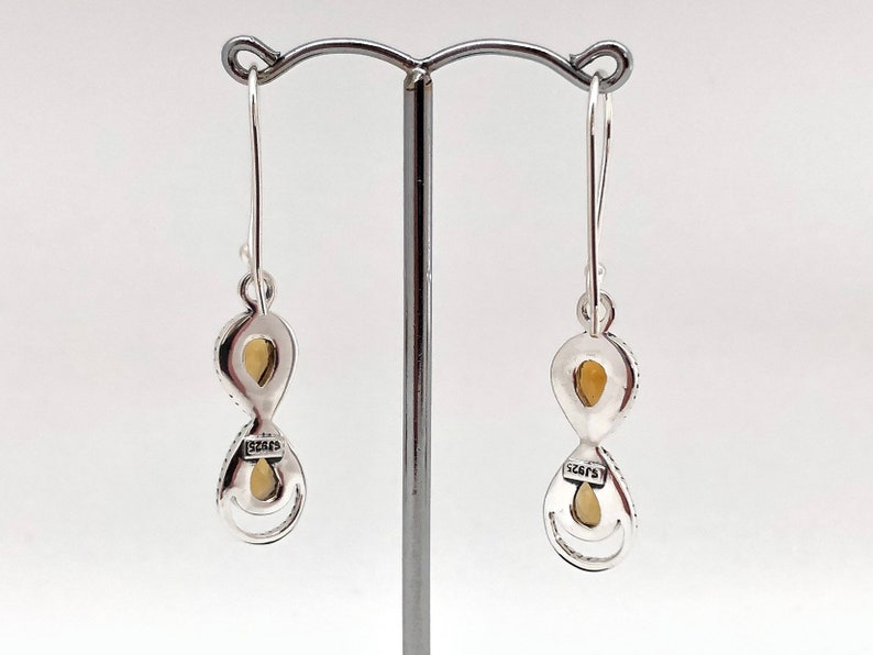 May include: A pair of silver drop earrings featuring two stacked, teardrop-shaped settings with light yellow stones. Each earring has a hook wire and a small rectangular plaque stamped with the text 925, indicating sterling silver composition.