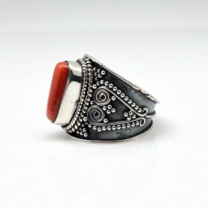 May include: A silver ring with an orange coral stone set in a silver bezel. The ring band is decorated with intricate silver filigree and beading.