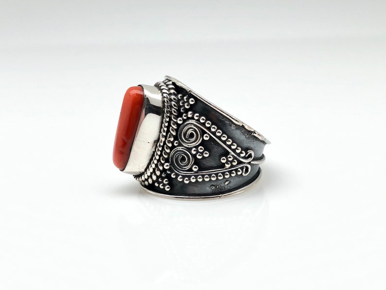 May include: A silver ring with an orange coral stone set in a silver bezel. The ring band is decorated with intricate silver filigree and beading.