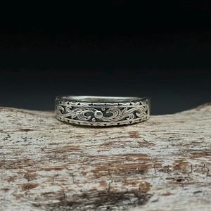 May include: A silver ring with an intricate, swirling floral pattern. The ring is approximately 0.25 inches wide and sits on a piece of weathered wood. The design features detailed scrollwork and small, raised dots.