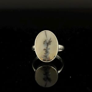 K&ouml;nnte beinhalten: Ovaler Ring mit einem Dendritenachat-Stein. Der Stein hat einen cremefarbenen Hintergrund mit schwarzen und grauen, baumartigen Einschl&uuml;ssen. Der Ring hat ein silberfarbenes Band und eine Fassung.