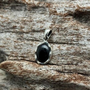 Puede incluir: Un colgante de plata con una piedra preciosa negra ovalada. El colgante tiene un dise&ntilde;o sencillo y elegante con un marco plateado pulido que rodea la piedra oscura. La argolla tambi&eacute;n es plateada.