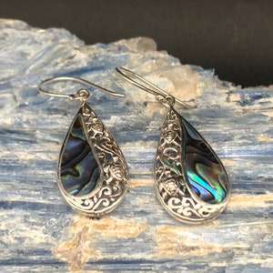 May include: A pair of silver dangle earrings with a teardrop-shaped abalone shell inlay. The earrings feature a delicate filigree design and a hook closure.