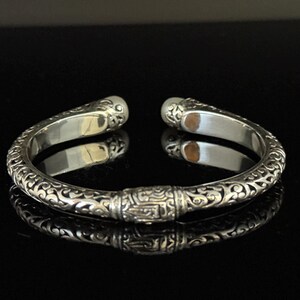 May include: Silver cuff bracelet with ornate, swirling patterns and a central rectangular design. The bracelet has rounded ends and a polished finish, reflecting the dark surface beneath.