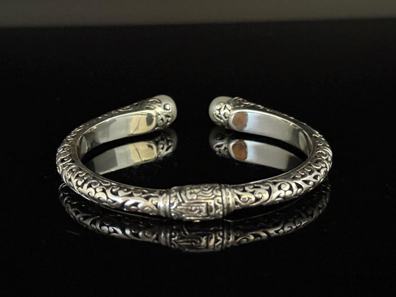 May include: Silver cuff bracelet with ornate, swirling patterns and a central rectangular design. The bracelet has rounded ends and a polished finish, reflecting the dark surface beneath.