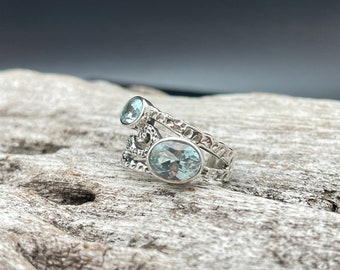 Puede incluir: Un anillo de plata con dos piedras preciosas de aguamarina azul claro. El anillo tiene una banda texturizada y un diseño delicado.