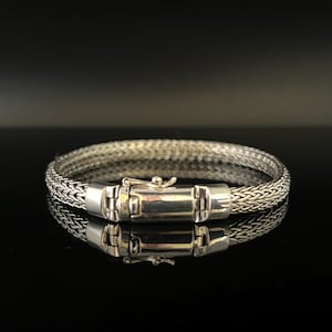 Puede incluir: Un brazalete de plata con un diseño de cadena tejida y un cierre de broche.
