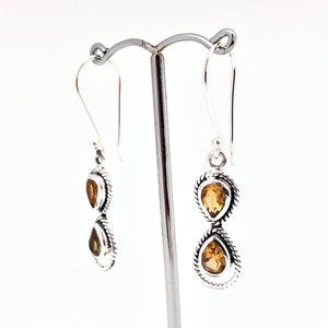 May include: A pair of silver drop earrings featuring two stacked teardrop-shaped citrine gemstones in each earring. The stones are set in twisted silver metal frames with hook-style ear wires, creating a vertical design with yellow-orange gems.