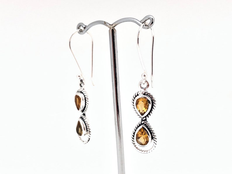 May include: A pair of silver drop earrings featuring two stacked teardrop-shaped citrine gemstones in each earring. The stones are set in twisted silver metal frames with hook-style ear wires, creating a vertical design with yellow-orange gems.