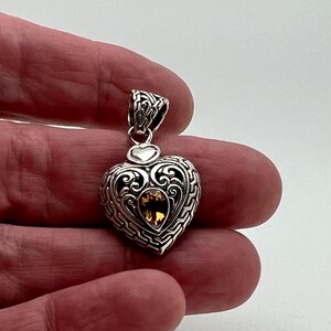 Puede incluir: Colgante de coraz&oacute;n de plata con un intrincado dise&ntilde;o de filigrana y una piedra preciosa de topacio dorado en el centro.